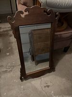 Full view of a rectangular mirror featuring a carved wooden frame with decorative top and bottom edges, standing on a concrete floor near furniture.