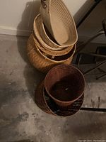 Photo showing six woven baskets stacked on a concrete floor against a wall. Visible oval beige basket on top, several round baskets below with light and dark tones.