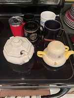 Top view of six beverage containers positioned on a stove, including ceramic teapots and various mugs and tumbler.