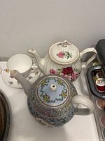 Photo of two teapots and one teacup with saucer placed on a white surface, showing floral designs and shapes.