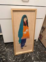 Framed textile art featuring a girl wearing a blue head covering, an orange tunic, and purple pants.