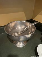 Overall view of pewter punch bowl with ladle inside, placed on a stove top.