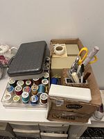 Photo of sewing thread spools in clear plastic organizers, desk caddy with scissors, measuring tape, and sewing tools, small boxes and tape inside a cardboard container.