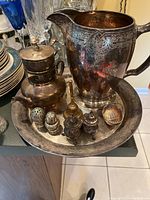 Photo showing large silver pitcher, teapot, serving tray, salt and pepper shakers, two small lidded containers, bell, and a small shell-shaped container arranged together.