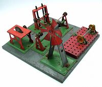 Full view of the Meccano steam engine accessories platform showing red and green painted metal components including a large windmill blade in the center, flywheels, and pulleys all mounted on a perforated gray base plate.