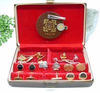 Top-down image of the open vintage case showing assorted cufflinks, tie bars, and tie tacks on red velvet lining with a wooden accessory piece labeled "TIE KLIPS".
