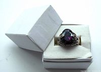 Ring inside a white jewelry box showing faceted deep purple stone with ornate silver mount.