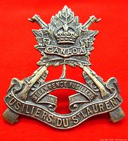 Front of the WWII era cap badge showing detailed design with maple leaf, rifles, crown and banners.