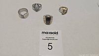 Four rings displayed on white surface with MaxSold tag number 5