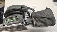 Side view of Genesis electric palm sander showing body, dust bag, and power cord coiled on top.