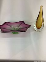 Photo showing side view of purple and green Murano glass bowl alongside amber teardrop vase on white background.
