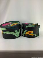 Side-by-side view of two round hat boxes covered in black fabric with colorful floral applique in pink, yellow, green, and dark blue. Each box has a matching vintage ladies hat inside or associated with it.