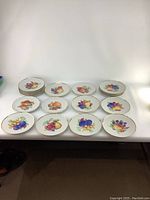 Front view of 14 vintage dessert plates laid out, each with different colorful fruit designs and gold trim on white ceramic surface.