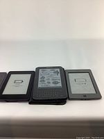 Three Kindle e-readers side by side on a white surface. Left and right devices show low battery indicators, center device displays black and white images of fish and aquatic creatures. The middle Kindle has a full QWERTY keyboard below the screen, the others have simpler button design.