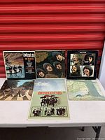 Seven Beatles record albums displayed on a white table against a red backdrop. Covers include Something New, Abbey Road, Let It Be, Rubber Soul, Beatles '65, and others.