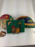 Three uniquely shaped and hand painted wooden place mats displayed upright on stands, showcasing abstract colorful designs including geometric shapes and nature imagery.