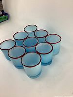 Nine hand made drinking glasses shown grouped together with frosted blue appearance and red rim on a white backdrop.