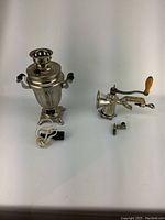 Photo showing vintage metal samovar with black handles and power cord on left, vintage manual kitchen grinder with wooden handle and clamp on right placed on white background.