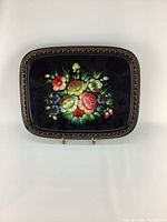 Top down view of the rectangular metal tray with black enamel base and colorful hand-painted floral design at the center, gold ornate border detail visible.