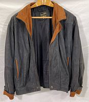 Full view of gray-black suede jacket with brown leather collar and pocket trim shown hanging on wooden hanger.