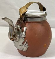 Front angled view showing the rounded clay body, metal handle with floral detail, milk glass lid set in metal rim, and decorative metal spout shaped like an animal climbing the body.