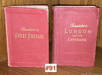 Two red cloth-bound Baedeker travel guide books side by side on a wooden shelf, titles embossed in gold: 'Great Britain' and 'London and Its Environs'