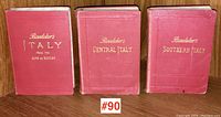 Three red hardbound antique Baedeker travel guides with gilt lettering on wooden bookshelf.