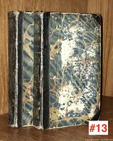 Two volumes of antiquarian books with marbled covers and black leather spines with gilt titles photographed upright on wooden shelf.