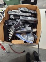 Photo showing an open box filled with multiple untested remote controls piled on top of each other.