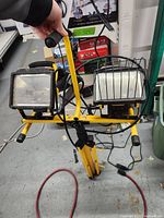 Front view of dual rectangular halogen lamps mounted on yellow tripod stand with handle grip. One lamp has a protective metal grid.