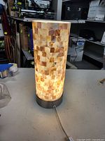 Cylindrical table lamp with mosaic square tiles lit up showing warm tones and a metallic base with a pull cord.