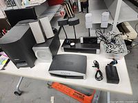 Photo showing a large assortment of Bose audio parts including subwoofers, satellite speakers, audio control unit, and cables on a table.