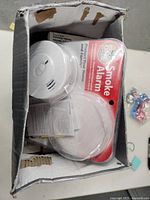 Photo of a Kidde Smoke Alarm partially unpacked in damaged cardboard box with instructions below it.