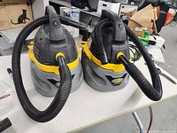 Two Stinger wet/dry vacuum cleaners side by side on a white table, with attached flexible black hoses and yellow handles.