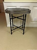 Black tray table with octagonal perforated metal tray top and cross-braced legs resembling bamboo style, against white paneled wall.