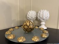 Glass serving tray with gold artichoke decorations and central gold artichoke piece in front, and two white artichoke pedestal statues behind