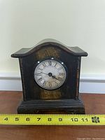 Wooden mantel clock with Roman numeral face, showing front and base with ruler for size reference.