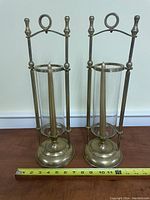 Pair of tall brass candle holders with glass cylinder holders and decorative loop handles on top shown against a white wall and wooden surface with measuring tape for scale
