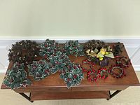 Wooden table with assorted Christmas decorations including artificial holly branches, two wreaths with pinecones and yellow ornaments, and small red bead decorations.