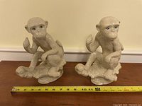Two beige ceramic monkey statues placed side by side with measuring tape showing length approximately 10 inches