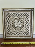 Close-up of a square needlework art piece displaying a complex geometric pattern in neutral tones within a gold frame, measuring approximately 12 inches wide.