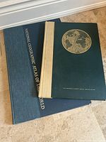 Photo of two vintage atlases stacked on floor showing their closed covers; one has green cover with beige spine and gold embossed globe illustration, the other has dark blue cloth cover with gold text on spine