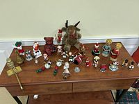 Full view of all vintage Christmas themed ornaments and trinkets on wooden table, showing diverse variety including plush reindeer and red bear, small Santa figurines, bell ornaments, and other holiday characters.
