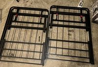Two black metal bed frames laid flat on a carpeted floor showing the grid structure and corner clips.
