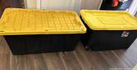 Photo showing two large black plastic storage containers with yellow lids; one container has wheels and the other does not.