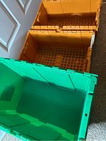 Three plastic logistic boxes placed on the floor near a door. Two yellow boxes are stacked and one green box is open showing the interior grid bottom.