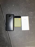 Photo showing three portable chargers side by side on a dark surface: large black, medium olive green, and small white