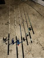 Six fishing rods laid out on a floor, five with spinning reels attached, various colors and lengths visible.