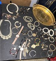 Photo of assorted costume jewelry on table including necklaces, bracelets, watches, earrings, and decorative tray.