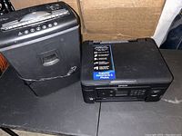 Image shows Epson XP-4100 printer next to a black paper shredder on a table, both with some usage wear.